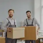 zwei Mitarbeiter von Umzugsunternehmen, die Kisten halten und in die Kamera schauen